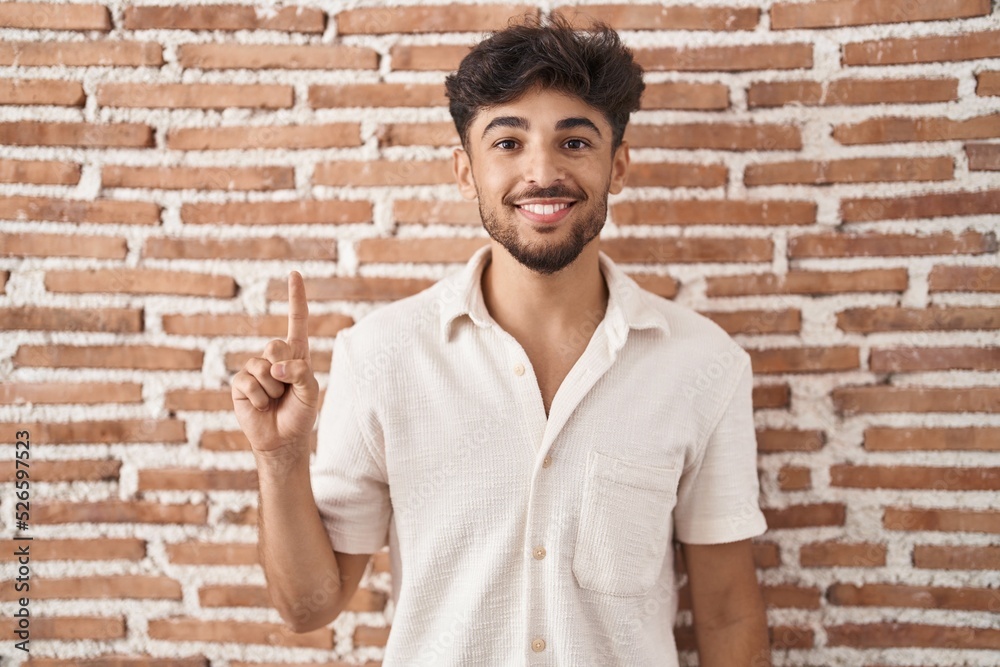 Arab man with beard standing over bricks wall background showing and pointing up with finger number one while smiling confident and happy. Arab man with beard standing over bricks wall background showing and pointing up with finger number one while smiling confident and happy.