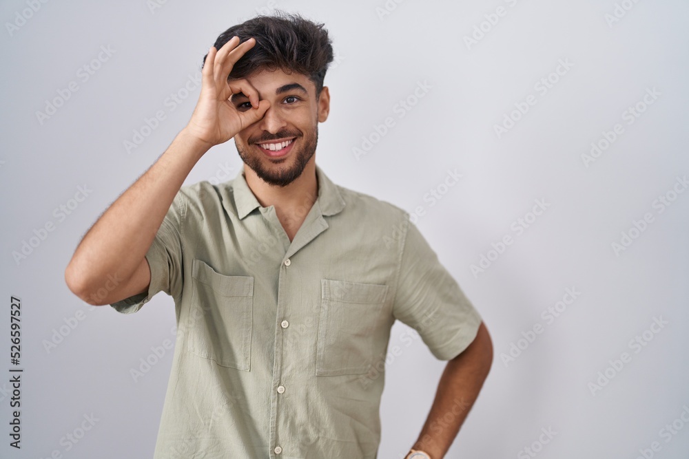 Arab man with beard standing over white background doing ok gesture with hand smiling, eye looking through fingers with happy face. Arab man with beard standing over white background doing ok gesture with hand smiling, eye looking through fingers with happy face.