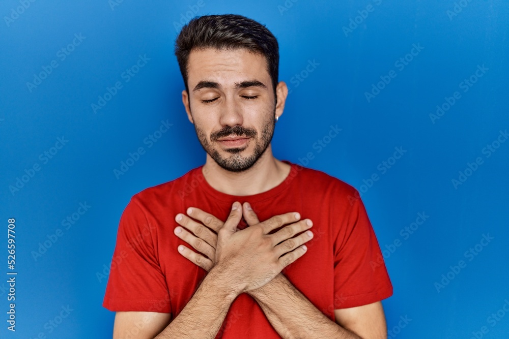 Young hispanic man with beard wearing red t shirt over blue background smiling with hands on chest with closed eyes and grateful gesture on face. health concept. Young hispanic man with beard wearing red t shirt over blue background smiling with hands on chest with closed eyes and grateful gesture on face. health concept.