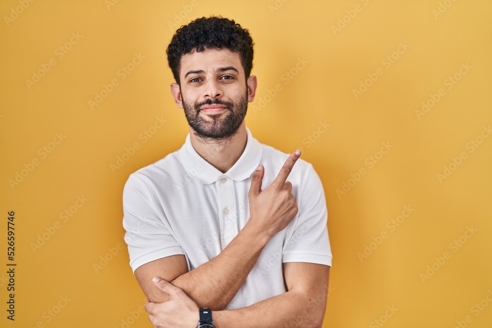 Arab man standing over yellow background pointing with hand finger to the side showing advertisement, serious and calm face