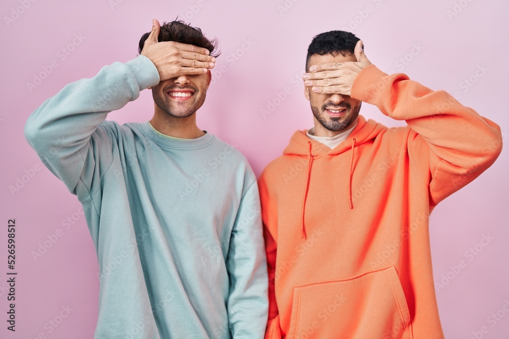 Young hispanic gay couple standing over pink background smiling and laughing with hand on face covering eyes for surprise. blind concept.