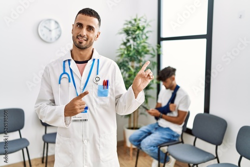 Hispanic doctor man at waiting room with pacient with arm injury smiling and looking at the camera pointing with two hands and fingers to the side.
