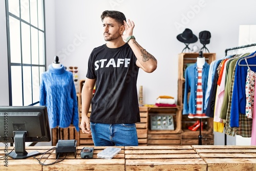 Young hispanic man working at retail boutique smiling with hand over ear listening an hearing to rumor or gossip. deafness concept.