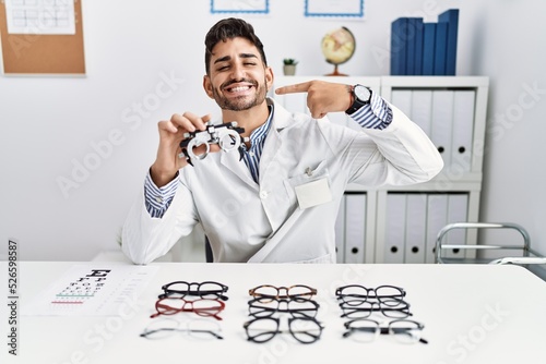 Young optician man holding optometry glasses smiling cheerful showing and pointing with fingers teeth and mouth. dental health concept.