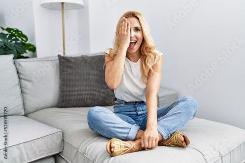 Beautiful blonde woman sitting on the sofa at home covering one eye with hand, confident smile on face and surprise emotion.