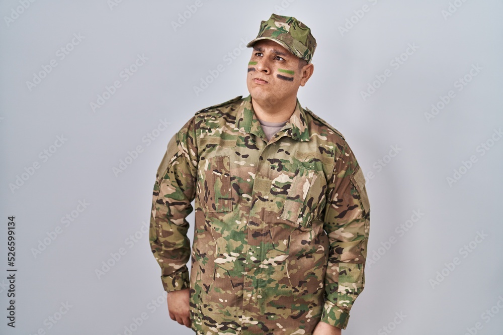 Hispanic young man wearing camouflage army uniform smiling looking to the side and staring away thinking. Hispanic young man wearing camouflage army uniform smiling looking to the side and staring away thinking.