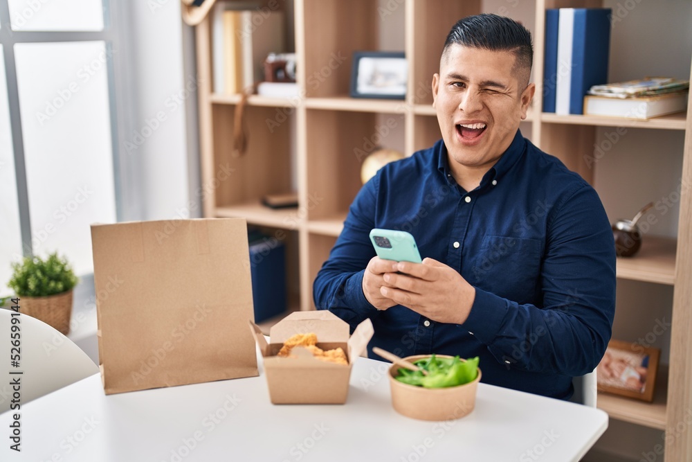 Hispanic young man eating take away food using smartphone winking looking at the camera with sexy expression, cheerful and happy face. Hispanic young man eating take away food using smartphone winking looking at the camera with sexy expression, cheerful and happy face.