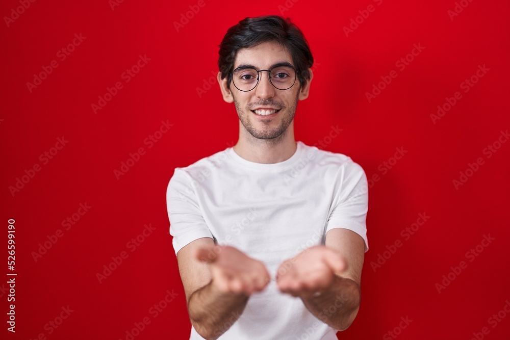Naklejka premium Young hispanic man standing over red background smiling with hands palms together receiving or giving gesture. hold and protection