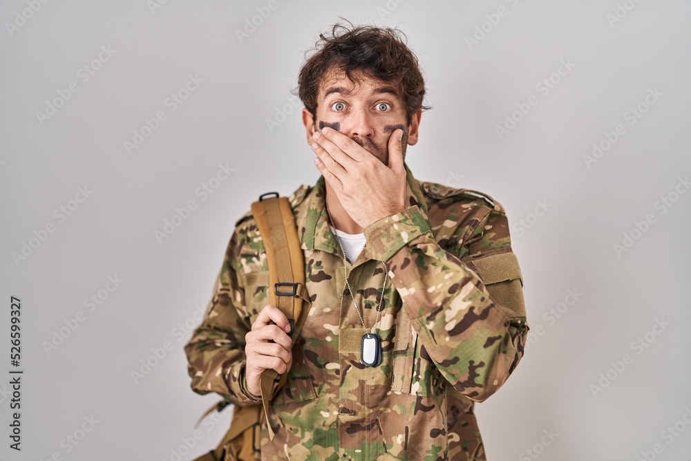 Hispanic young man wearing camouflage army uniform shocked covering mouth with hands for mistake. secret concept. Hispanic young man wearing camouflage army uniform shocked covering mouth with hands for mistake. secret concept.