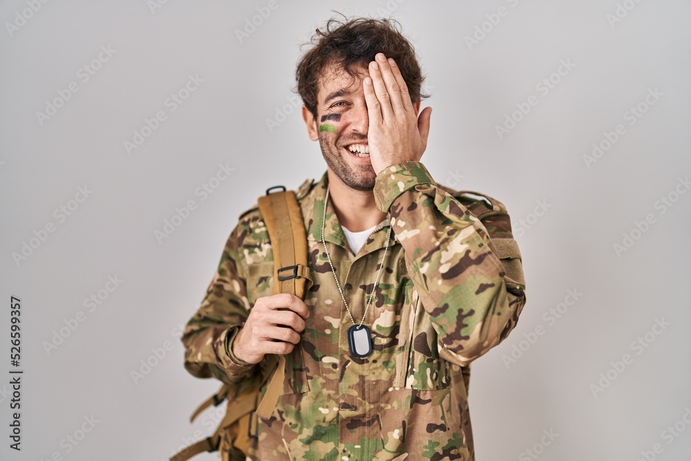 Hispanic young man wearing camouflage army uniform covering one eye with hand, confident smile on face and surprise emotion.