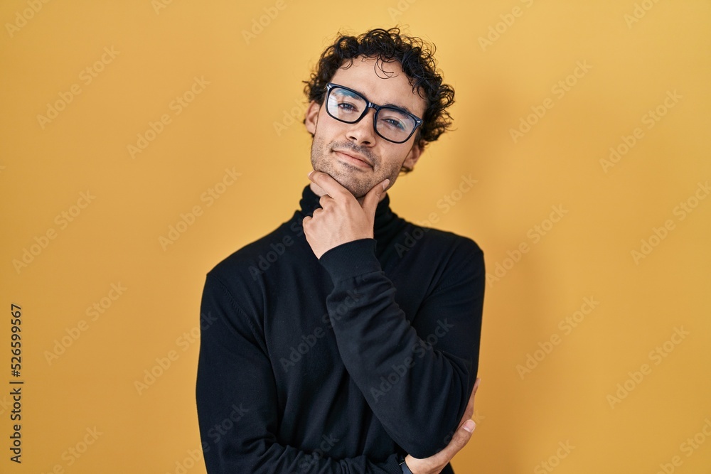 Hispanic man standing over yellow background looking confident at the camera smiling with crossed arms and hand raised on chin. thinking positive. Hispanic man standing over yellow background looking confident at the camera smiling with crossed arms and hand raised on chin. thinking positive.