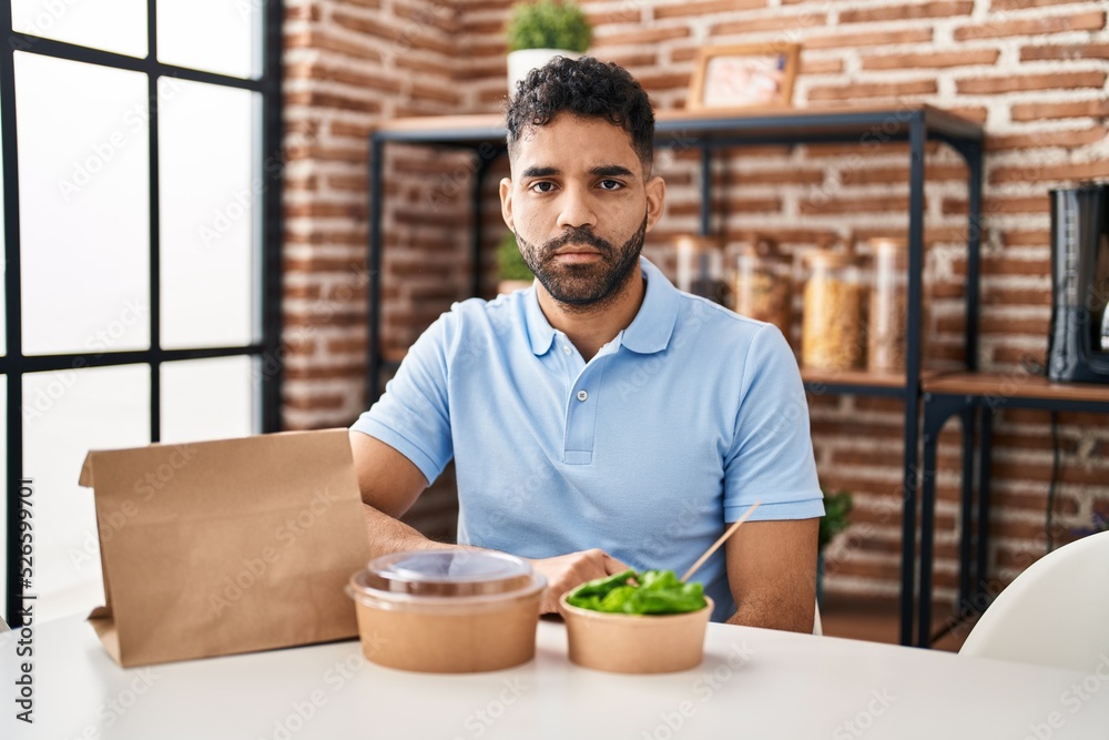 Hispanic man with beard eating delivery salad thinking attitude and sober expression looking self confident Hispanic man with beard eating delivery salad thinking attitude and sober expression looking self confident