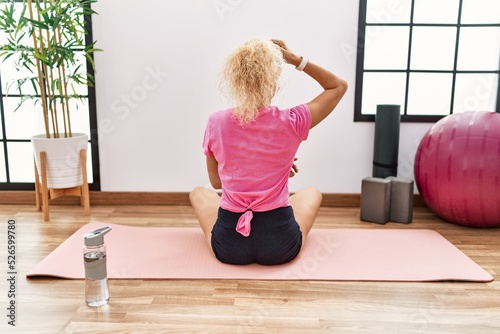 Middle age blonde woman sitting on yoga mat backwards thinking about doubt with hand on head