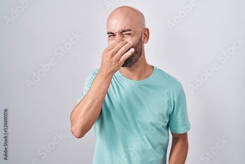 Middle age bald man standing over white background smelling something stinky and disgusting, intolerable smell, holding breath with fingers on nose. bad smell