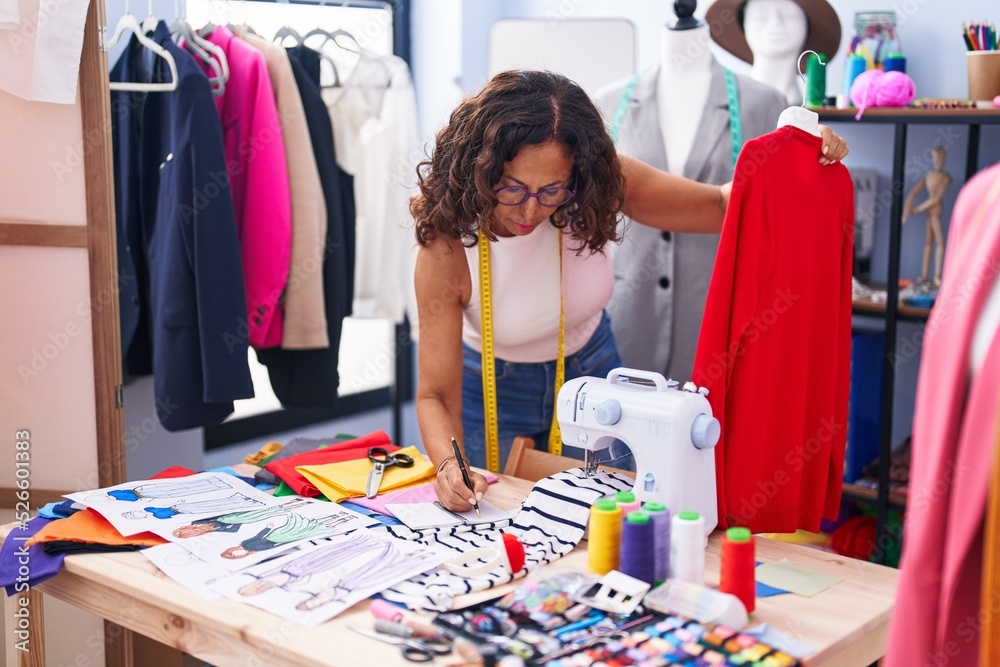Fototapeta premium Middle age woman tailor holding t shirt writing on notebook at clothing factory