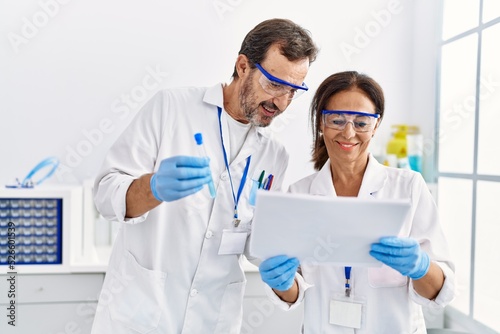 Middle age man and woman partners wearing scientist uniform reading document and holding test tube at laboratory