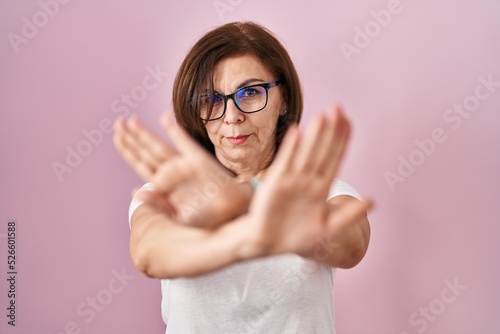 Middle age hispanic woman standing over pink background rejection expression crossing arms and palms doing negative sign  angry face