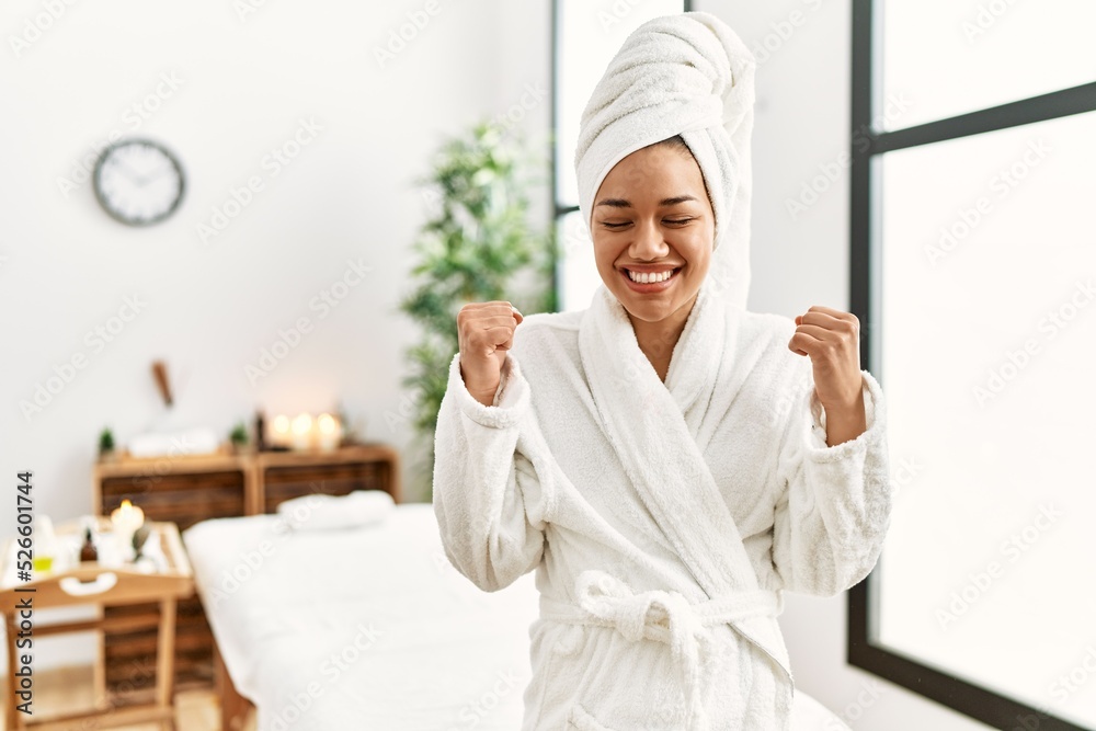 Young brunette woman wearing towel and bathrobe standing at beauty center excited for success with arms raised and eyes closed celebrating victory smiling. winner concept. Young brunette woman wearing towel and bathrobe standing at beauty center excited for success with arms raised and eyes closed celebrating victory smiling. winner concept.
