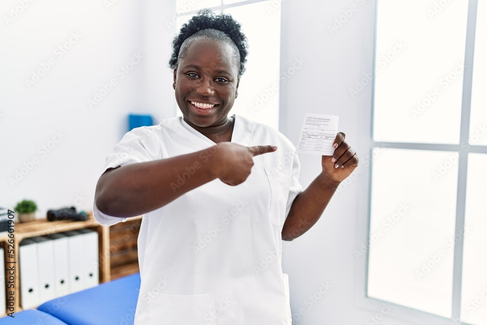 Young african woman holding covid record card at clinic smiling happy pointing with hand and finger
