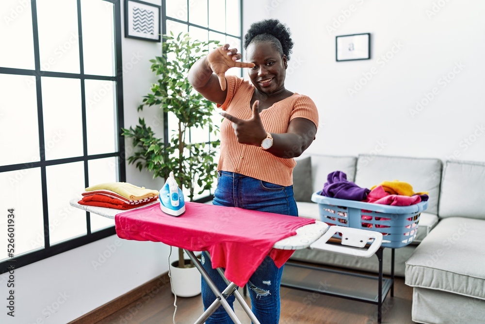 Obraz premium Young african woman ironing clothes at home smiling making frame with hands and fingers with happy face. creativity and photography concept.