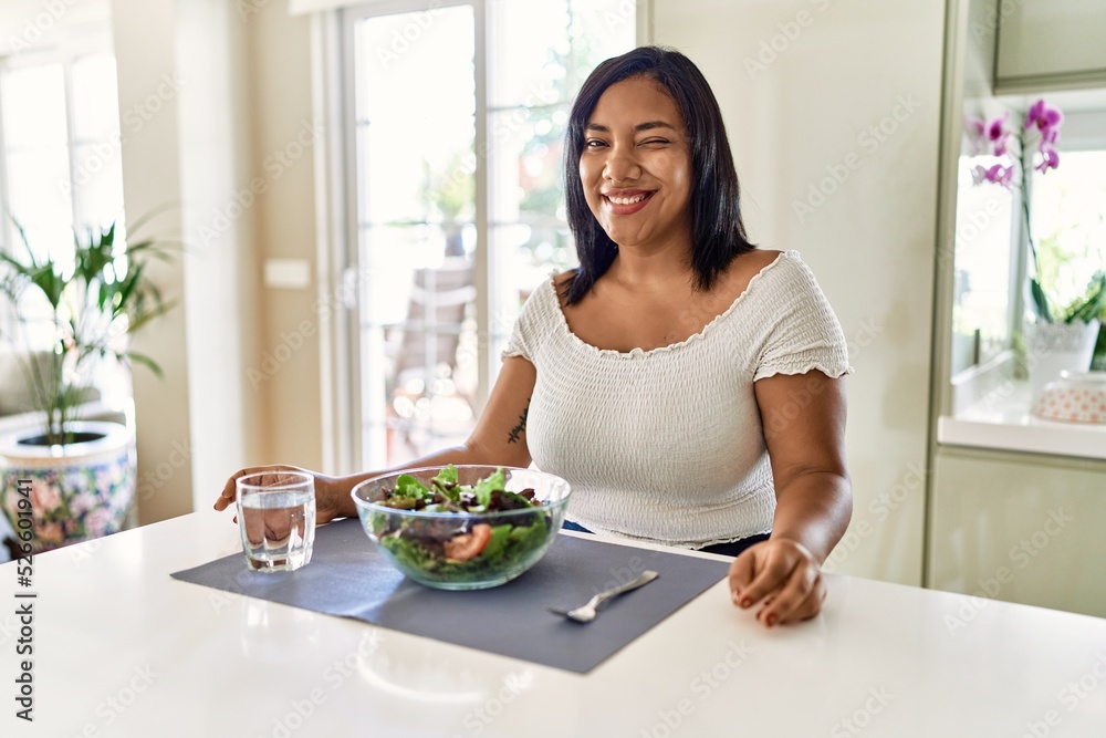 Young hispanic woman eating healthy salad at home winking looking at the camera with sexy expression, cheerful and happy face.