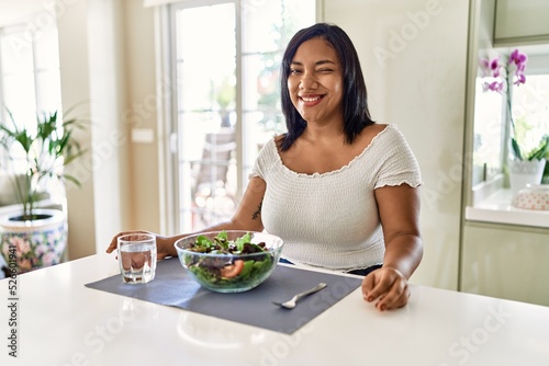 Young hispanic woman eating healthy salad at home winking looking at the camera with sexy expression  cheerful and happy face.