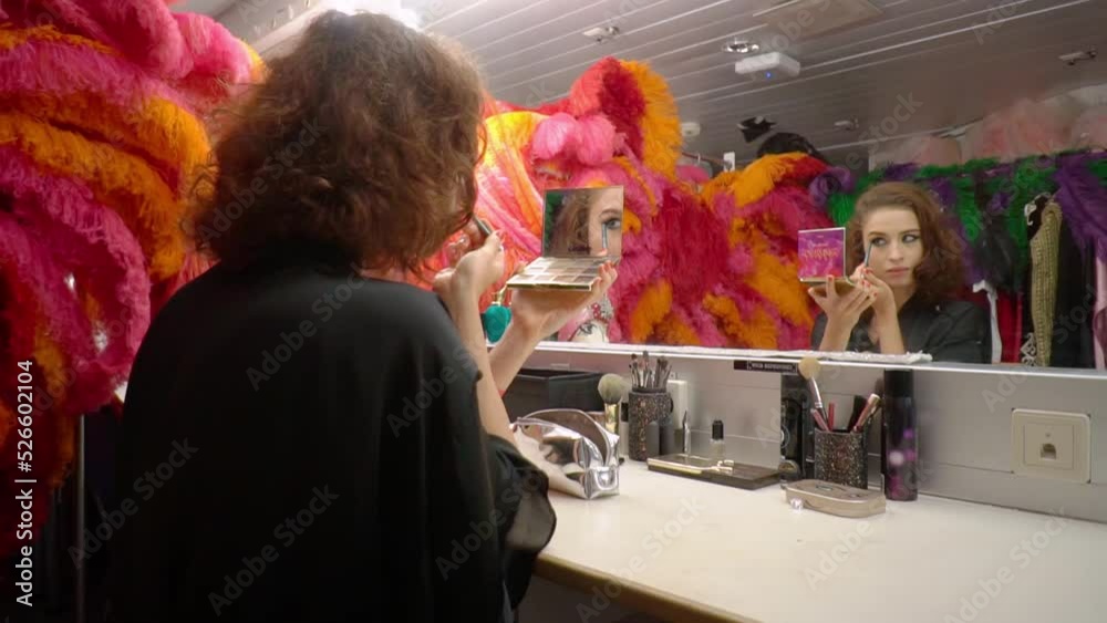 Beautiful woman actress applies eye makeup,holds compact eye shadow palette with mirror in front of her,sits in dressing room at table. Rear side view,camera zooming.Bright dance costumes,feathers.
