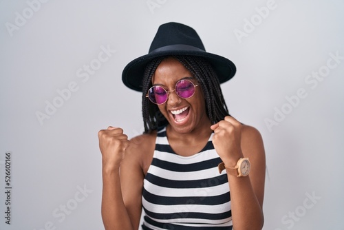 Young african american with braids wearing hat and sunglasses very happy and excited doing winner gesture with arms raised, smiling and screaming for success. celebration concept.