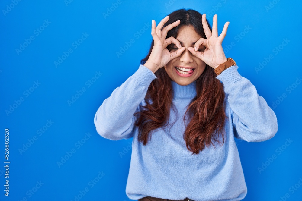 Hispanic young woman standing over blue background doing ok gesture like binoculars sticking tongue out, eyes looking through fingers. crazy expression. Hispanic young woman standing over blue background doing ok gesture like binoculars sticking tongue out, eyes looking through fingers. crazy expression.
