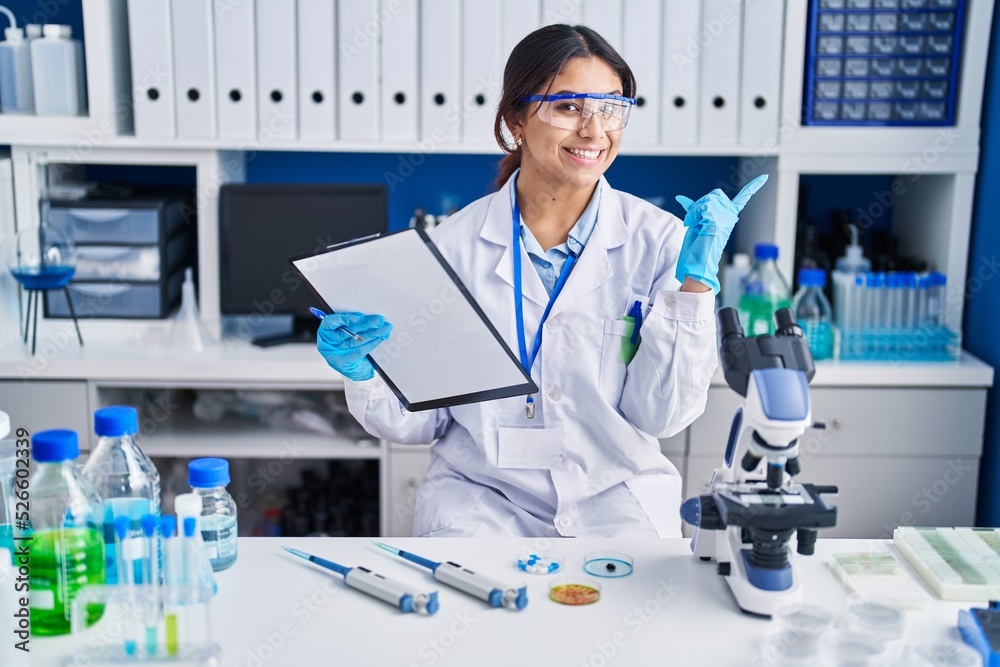 Hispanic young woman working at scientist laboratory with a big smile on face, pointing with hand finger to the side looking at the camera.