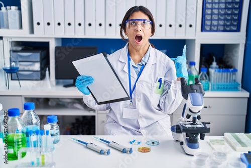 Hispanic young woman working at scientist laboratory amazed and surprised looking up and pointing with fingers and raised arms.