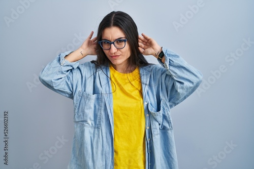 Young hispanic woman standing over blue background trying to hear both hands on ear gesture, curious for gossip. hearing problem, deaf