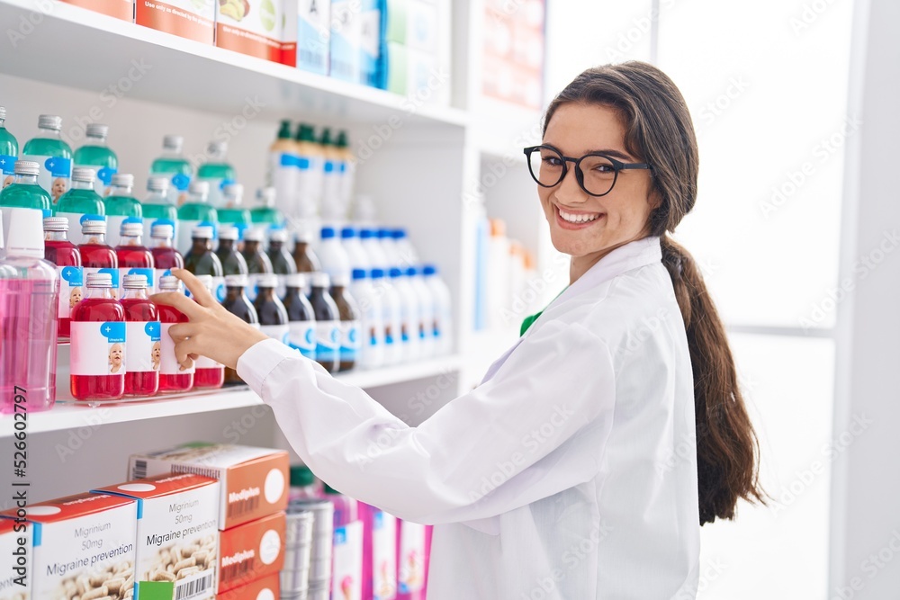 Young hispanic woman pharmacist smiling confident organizing shelving at pharmacy