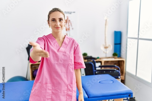 Young blonde woman working at pain recovery clinic smiling friendly offering handshake as greeting and welcoming. successful business.