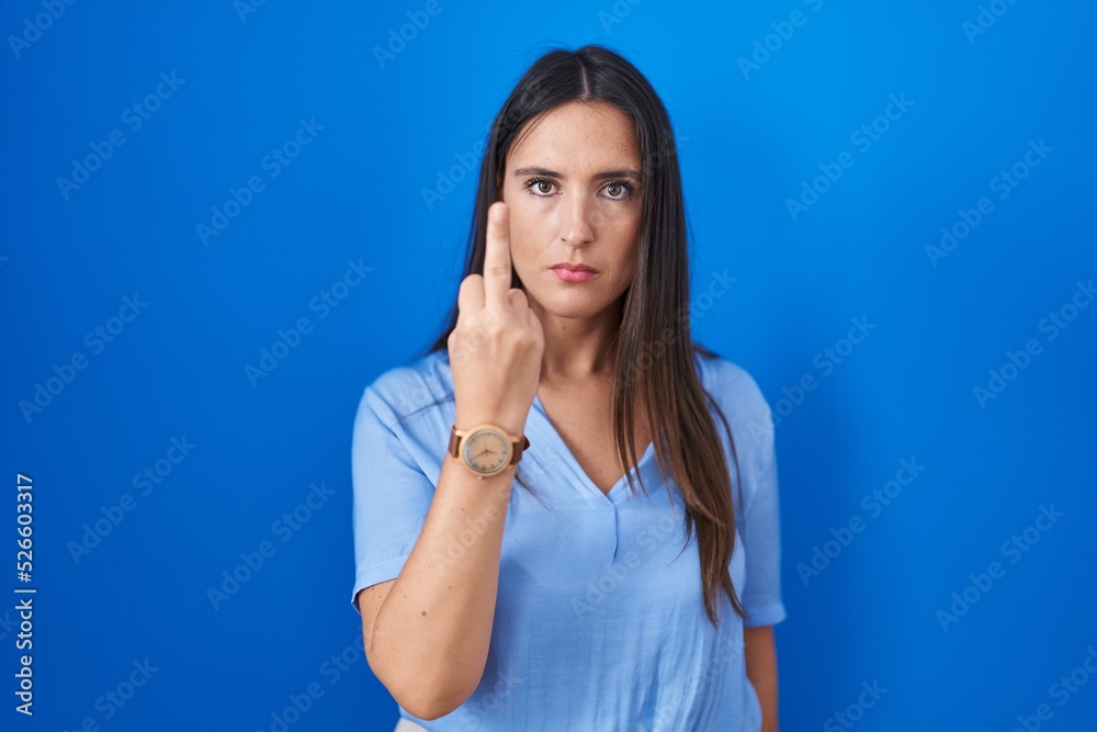 Young brunette woman standing over blue background showing middle finger, impolite and rude fuck off expression Young brunette woman standing over blue background showing middle finger, impolite and rude fuck off expression