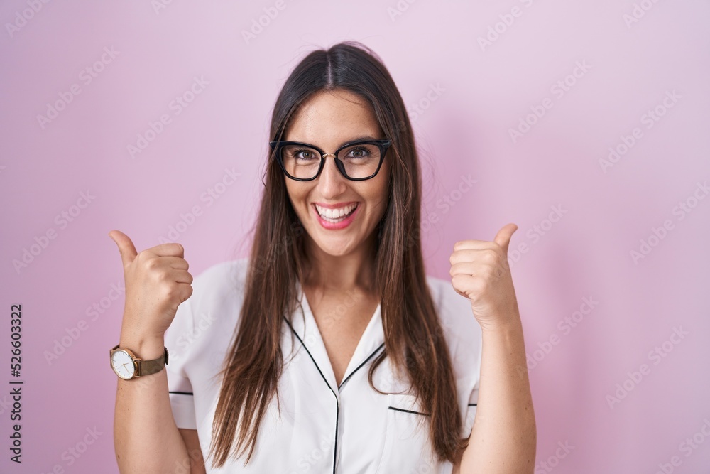Young brunette woman wearing glasses standing over pink background success sign doing positive gesture with hand, thumbs up smiling and happy. cheerful expression and winner gesture.