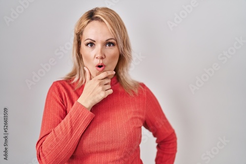 Blonde woman standing over isolated background looking fascinated with disbelief, surprise and amazed expression with hands on chin