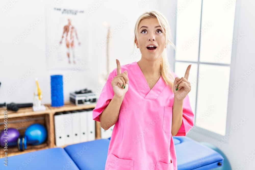 Young caucasian woman working at pain recovery clinic amazed and surprised looking up and pointing with fingers and raised arms. Young caucasian woman working at pain recovery clinic amazed and surprised looking up and pointing with fingers and raised arms.