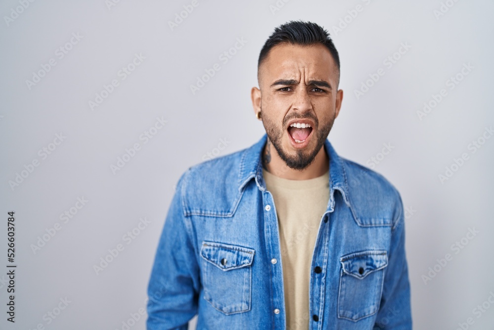 Young hispanic man standing over isolated background angry and mad screaming frustrated and furious, shouting with anger. rage and aggressive concept. Young hispanic man standing over isolated background angry and mad screaming frustrated and furious, shouting with anger. rage and aggressive concept.