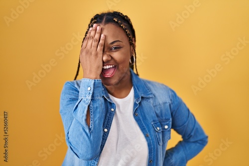 African american woman with braids standing over yellow background covering one eye with hand, confident smile on face and surprise emotion.