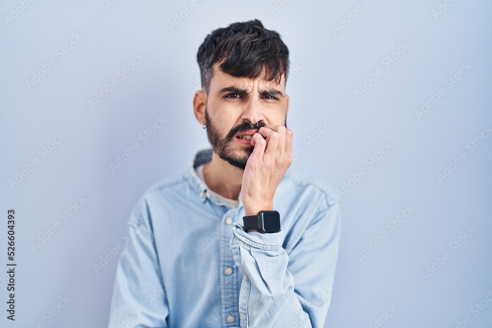 Young hispanic man with beard standing over blue background looking stressed and nervous with hands on mouth biting nails. anxiety problem. Young hispanic man with beard standing over blue background looking stressed and nervous with hands on mouth biting nails. anxiety problem.