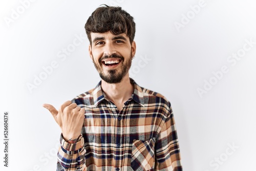 Hispanic man with beard standing over isolated background smiling with happy face looking and pointing to the side with thumb up.