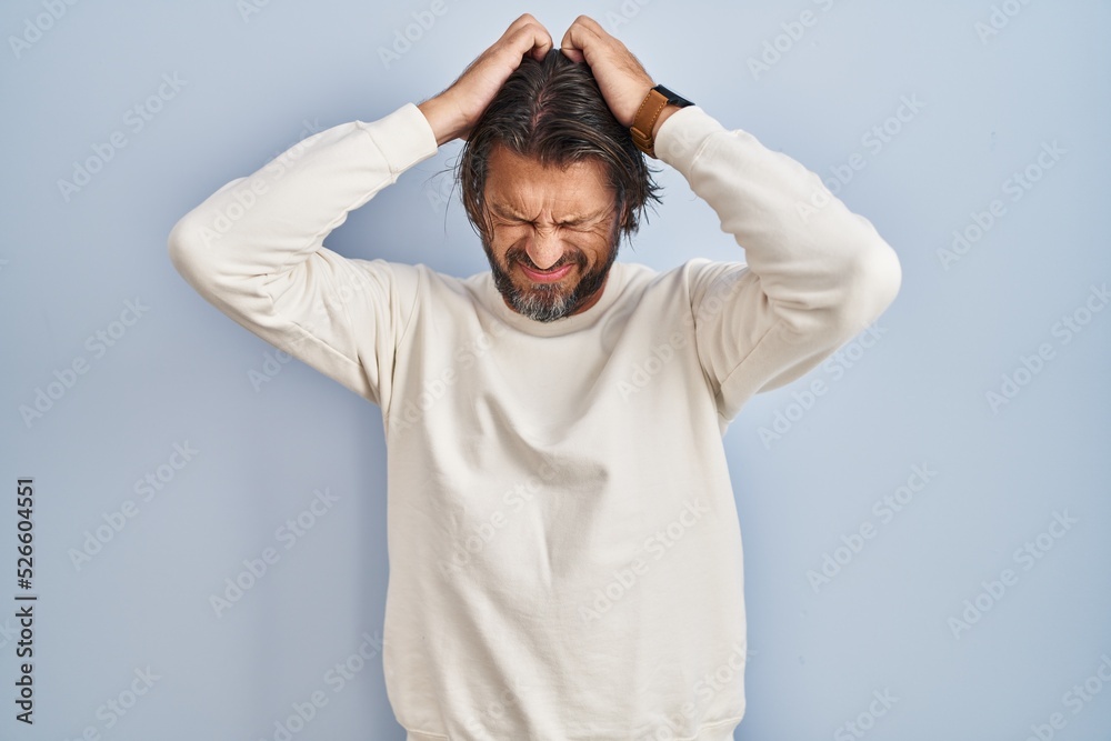 Handsome middle age man wearing casual sweater over blue background suffering from headache desperate and stressed because pain and migraine. hands on head. Handsome middle age man wearing casual sweater over blue background suffering from headache desperate and stressed because pain and migraine. hands on head.