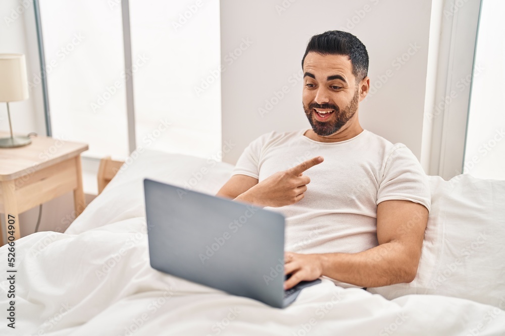 Young hispanic man with beard using computer laptop on the bed smiling happy pointing with hand and finger to the side Young hispanic man with beard using computer laptop on the bed smiling happy pointing with hand and finger to the side