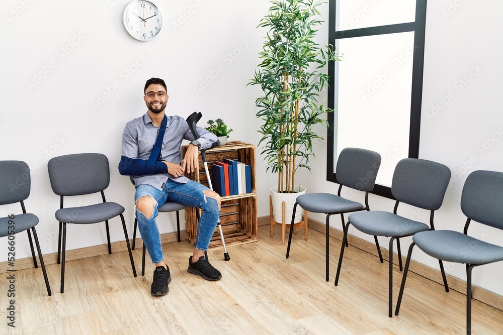 Young arab man smiling confident sitting on chair with arm sling holding crutch at clinic waiting room Young arab man smiling confident sitting on chair with arm sling holding crutch at clinic waiting room