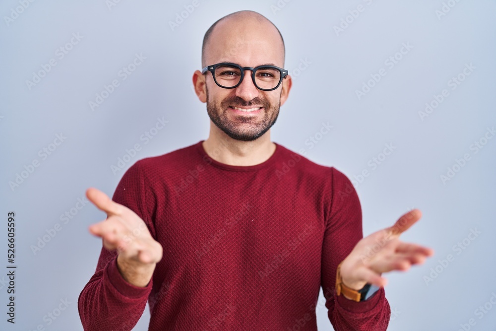 Young bald man with beard standing over white background wearing glasses smiling cheerful offering hands giving assistance and acceptance. Young bald man with beard standing over white background wearing glasses smiling cheerful offering hands giving assistance and acceptance.