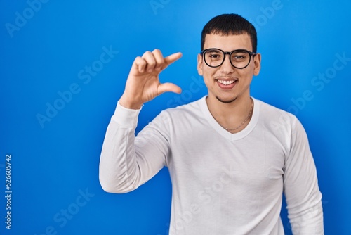 Young arab man wearing casual white shirt and glasses smiling and confident gesturing with hand doing small size sign with fingers looking and the camera. measure concept.