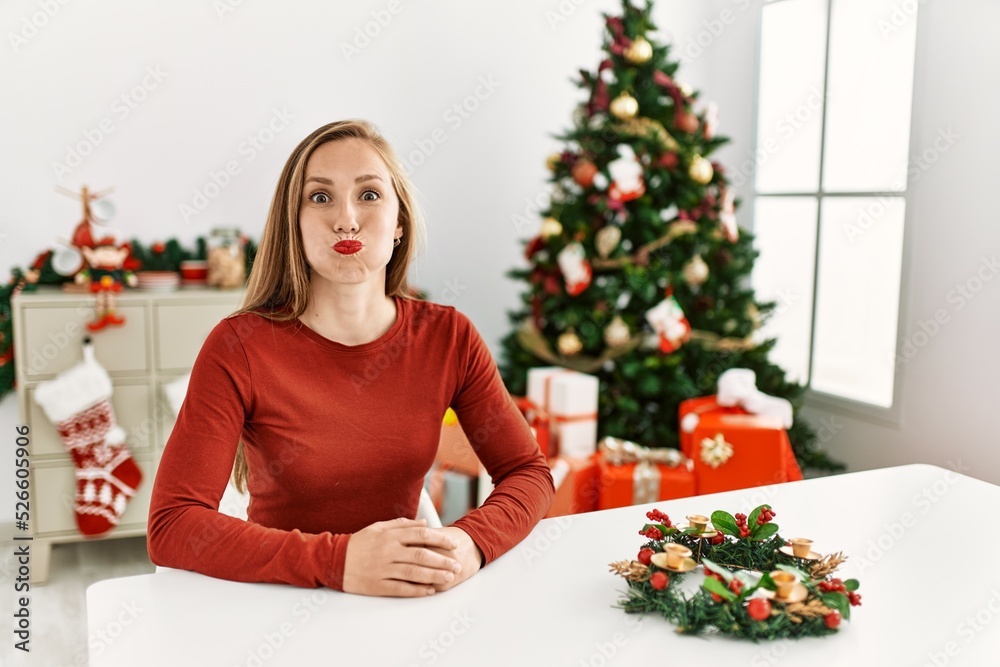 Caucasian young blonde woman sitting on the table by christmas tree puffing cheeks with funny face. mouth inflated with air, crazy expression. Caucasian young blonde woman sitting on the table by christmas tree puffing cheeks with funny face. mouth inflated with air, crazy expression.