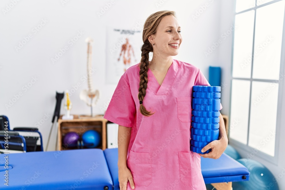 Young caucasian woman wearing physiotherapist uniform holding foam roller at physiotherapy clinic