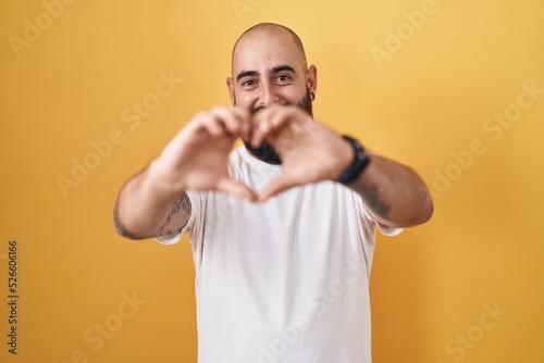 Young hispanic man with beard and tattoos standing over yellow background smiling in love doing heart symbol shape with hands. romantic concept.